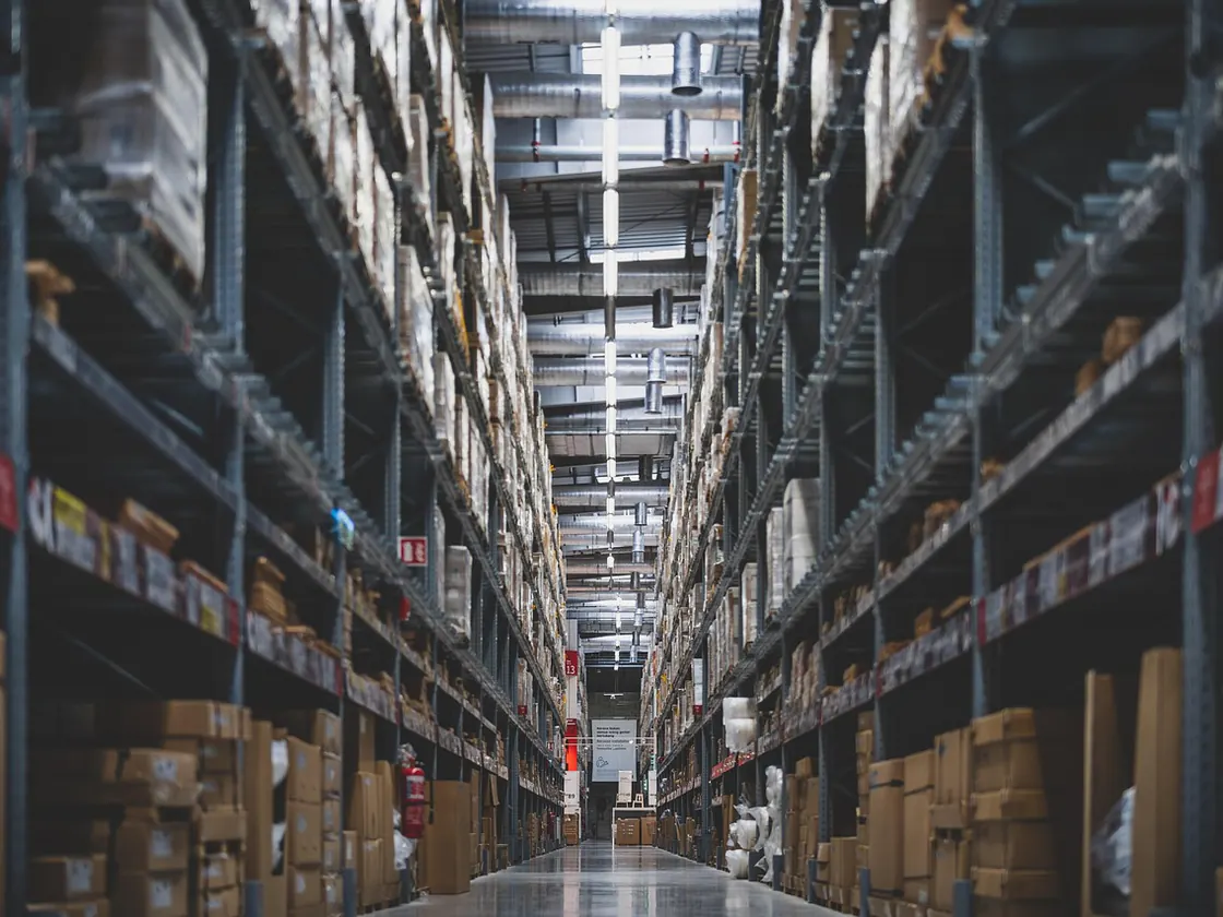 Long warehouse aisle lined with tall pallet racking stacked with cardboard cartons under bright overhead lights