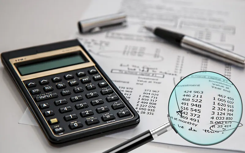 Calculator, pen and a printed sheet of figures under a magnifying glass on a desk